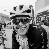 Un ciclista sorridente con una giacca da ciclismo nera con la scritta 'BIKE INDUSTRIES' sulla manica e un casco bianco, indossa occhiali da sole che riflettono l'ambiente.