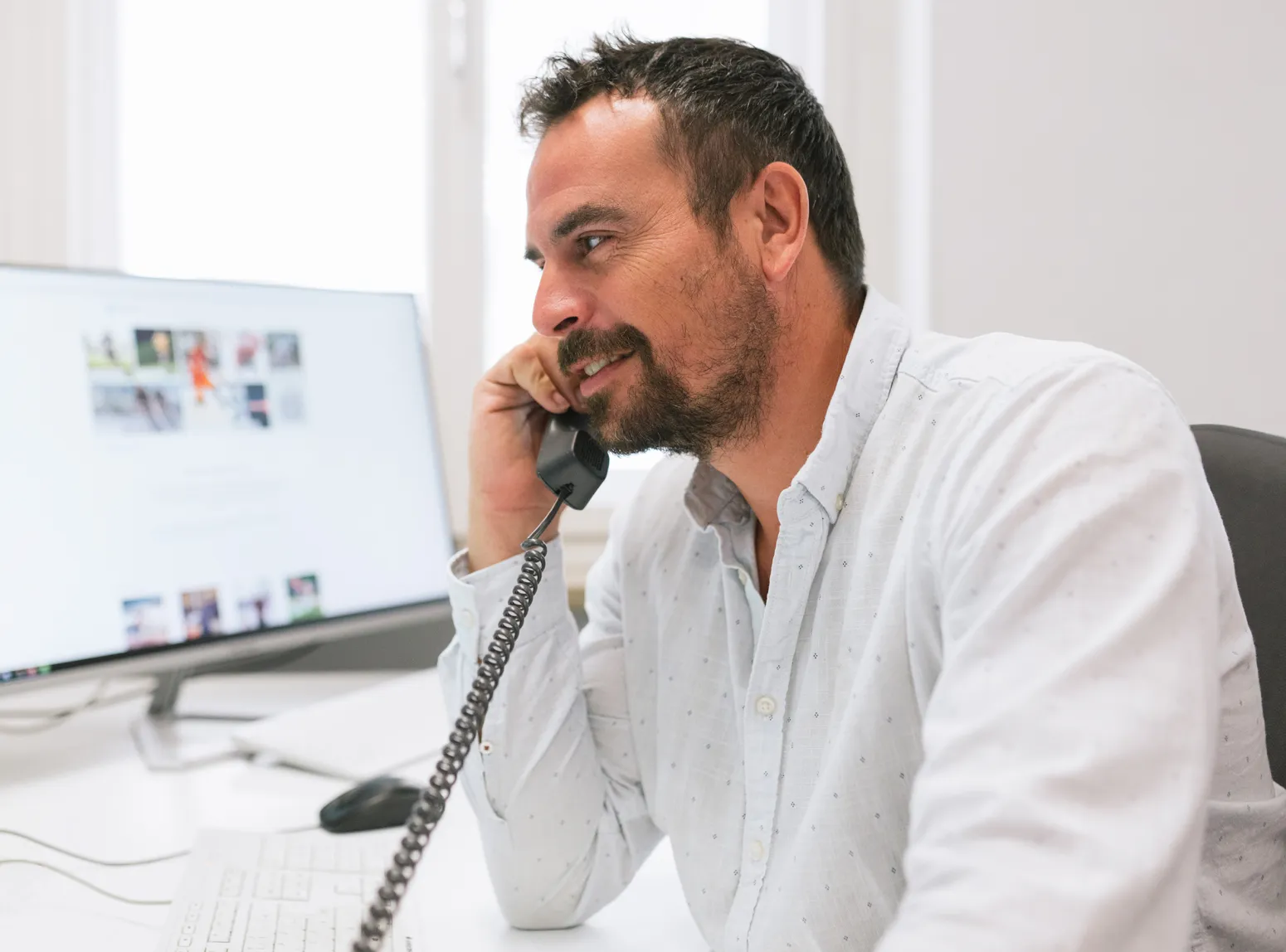 Un uomo con la barba è seduto a una scrivania davanti a un computer, sorridendo mentre parla al telefono con una vecchia cornetta.