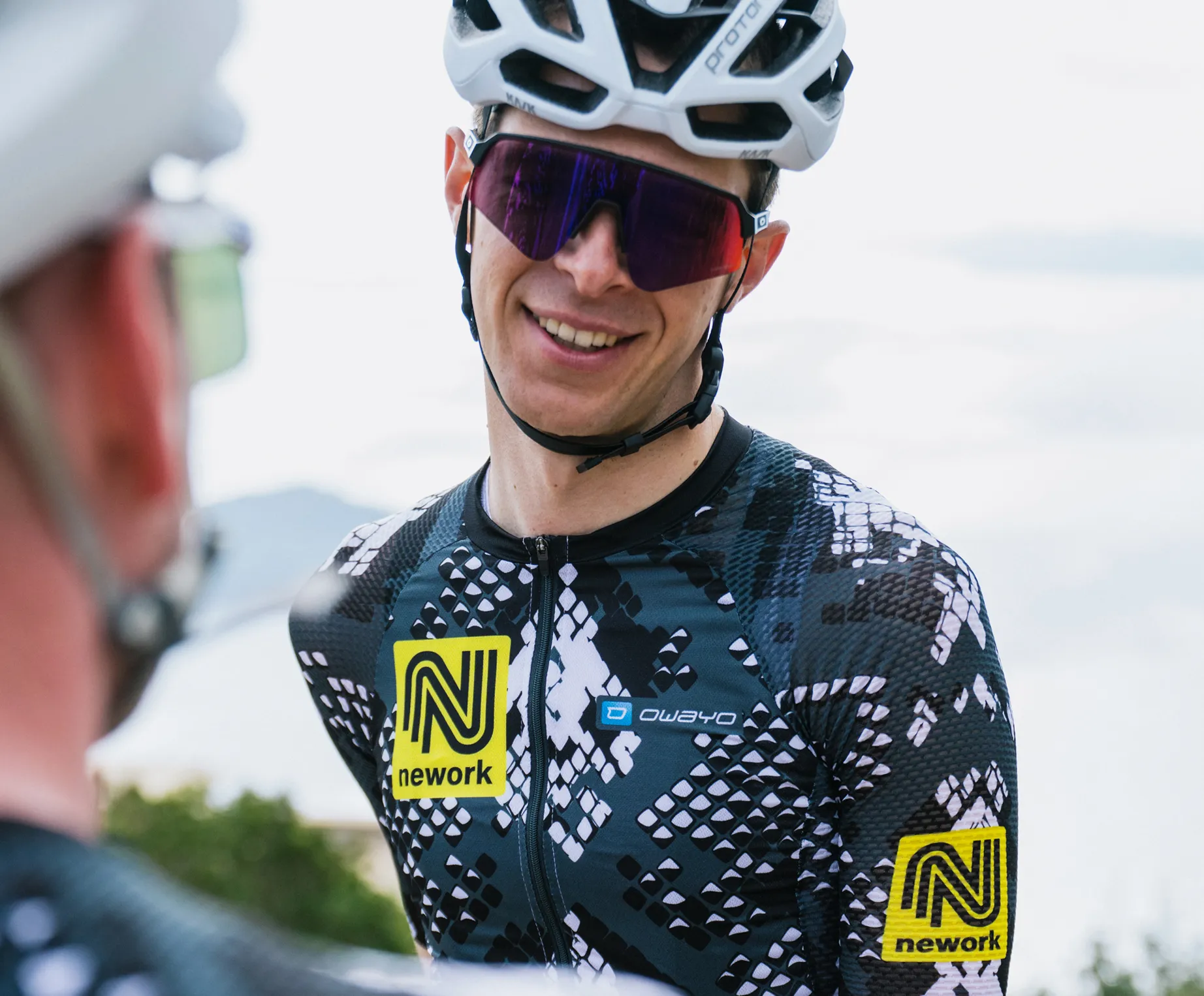 Un ciclista indossa una maglia personalizzata a motivi neri e bianchi con un logo giallo 'nework' e un logo 'owayo' sul petto.