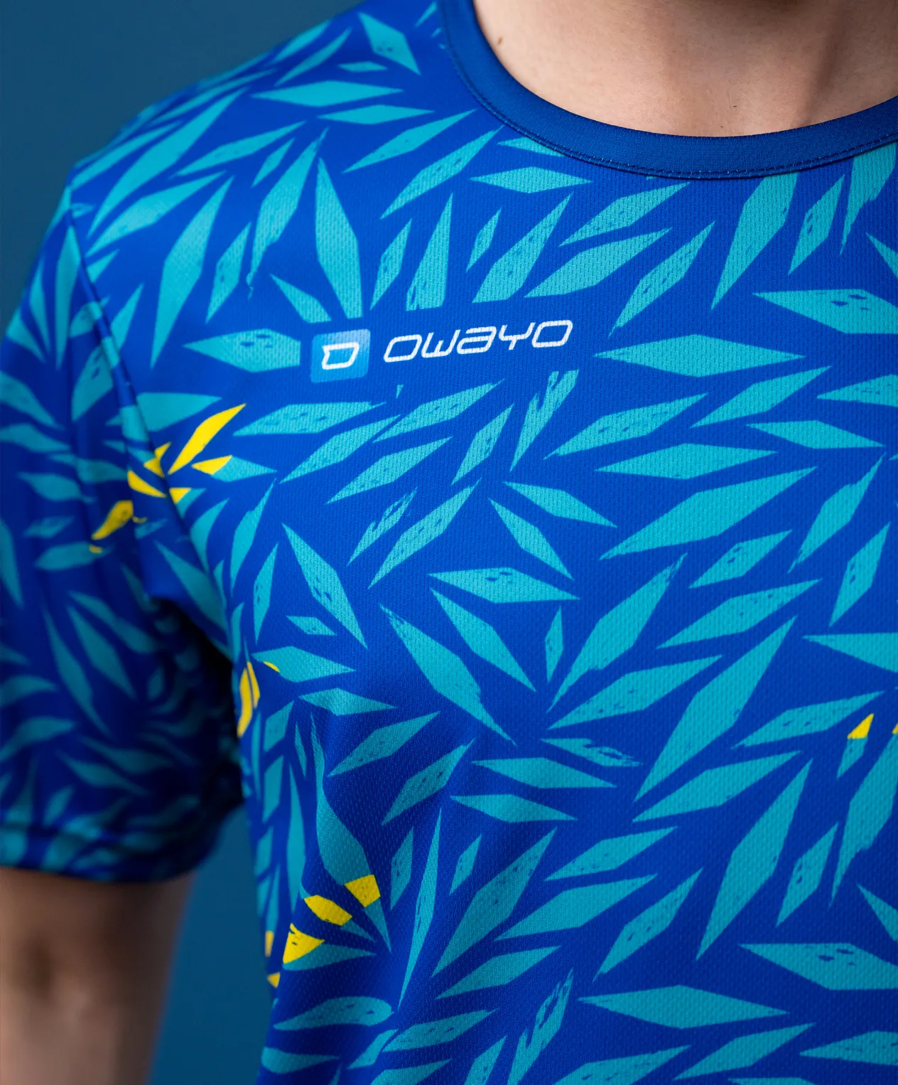 Una maglietta sportiva a maniche corte con un motivo blu a forma di foglia in varie tonalità di verde con accenti gialli.