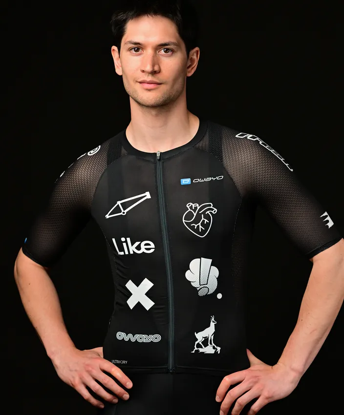 Un giovane indossa una maglietta sportiva nera aderente decorata con vari loghi e design bianchi, posando su uno sfondo nero.