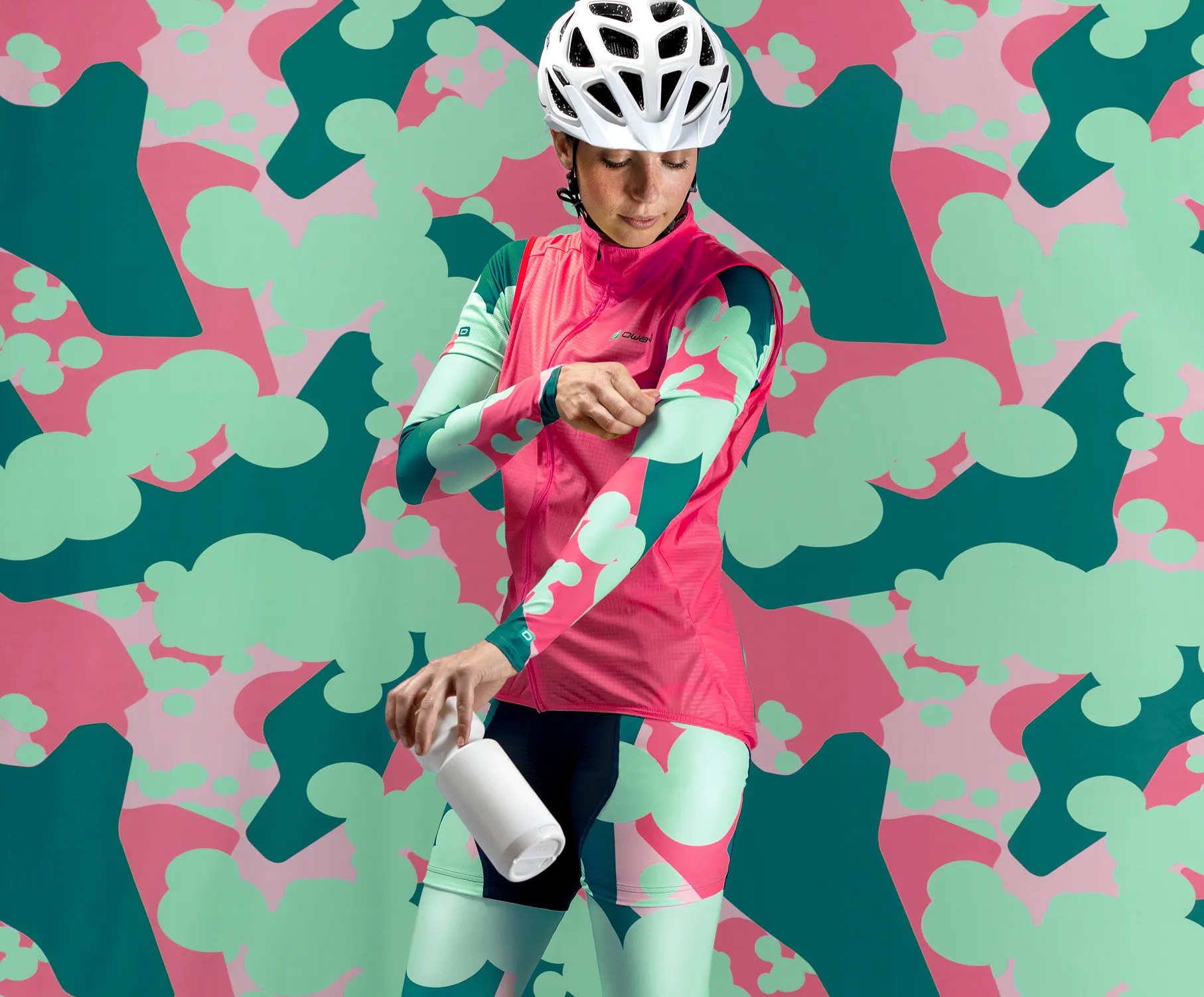 Una ciclista in abbigliamento sportivo personalizzato rosa e verde con un motivo astratto, che regola le maniche mentre tiene una bottiglia bianca, davanti a uno sfondo colorato con lo stesso motivo.