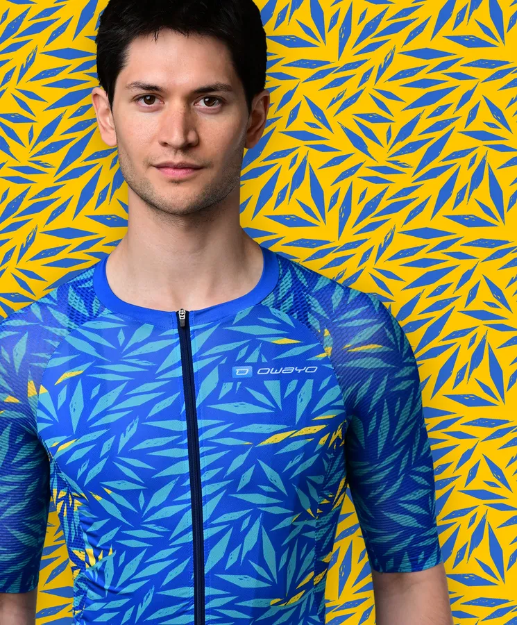 Un uomo indossa una maglia sportiva blu a maniche corte con un motivo di foglie blu e gialle che ricorda una pianta tropicale, davanti a uno sfondo giallo con motivi di foglie blu.
