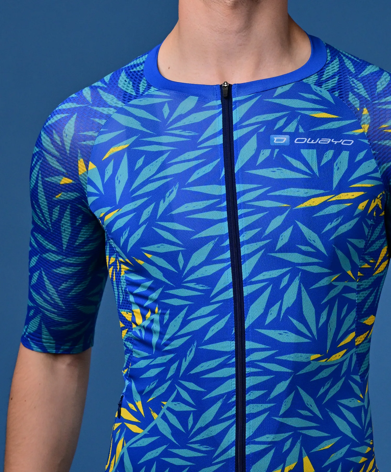 Un uomo indossa una maglia sportiva blu a maniche corte con un motivo tropicale di foglie in turchese e giallo e una cerniera frontale.