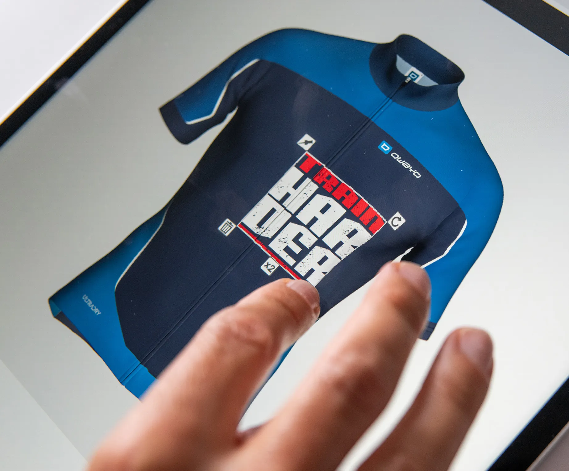 Una mano tocca uno schermo che mostra una maglia sportiva personalizzata blu scuro e blu con la scritta 'TRAIN HARDER' e il logo owayo.