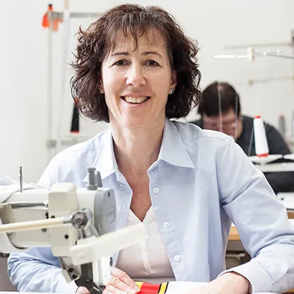 Una donna sorride alla telecamera mentre lavora a una macchina da cucire in un laboratorio, con un'altra persona visibile sullo sfondo che lavora anche a una macchina da cucire.
