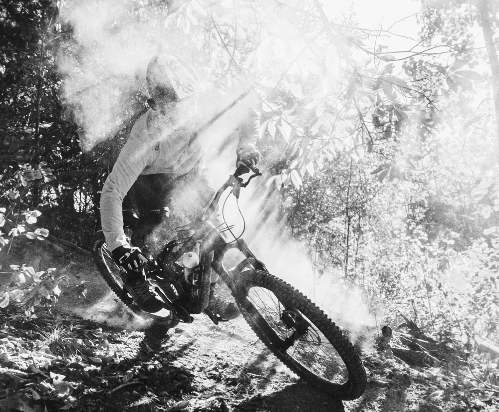 Un ciclista di montagna in movimento su un sentiero forestale, circondato da polvere e raggi di luce, indossa una maglia a maniche lunghe di owayo.