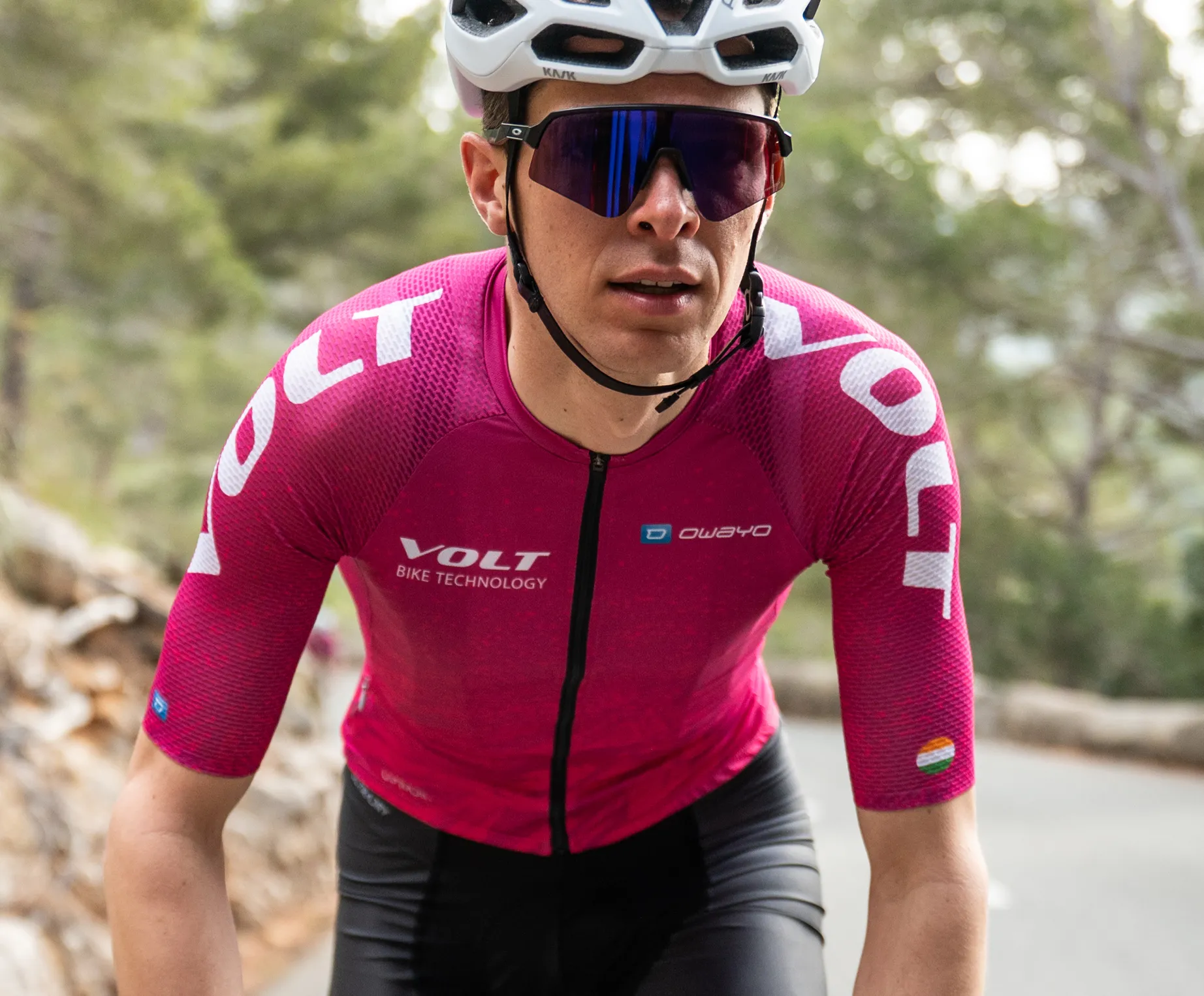 Un ciclista indossa una maglia da ciclismo rosa a maniche corte con il logo 'VOLT BIKE TECHNOLOGY' e il logo 'owayo' sul petto, che pedala su una strada in una zona boscosa.