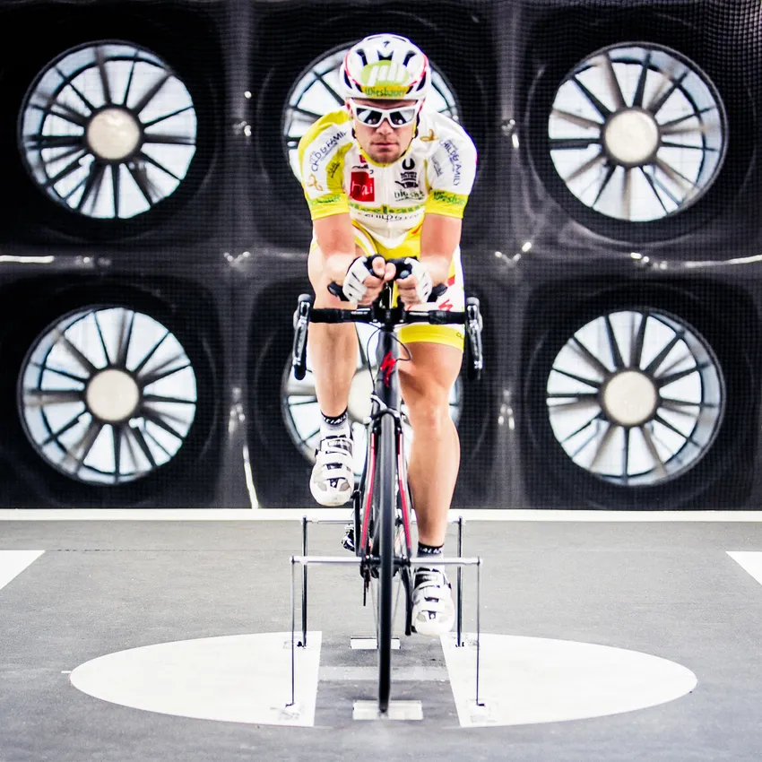 Un ciclista che si allena su una bicicletta statica in un laboratorio di test per ottimizzare l'aerodinamica, con grandi ventilatori sullo sfondo.