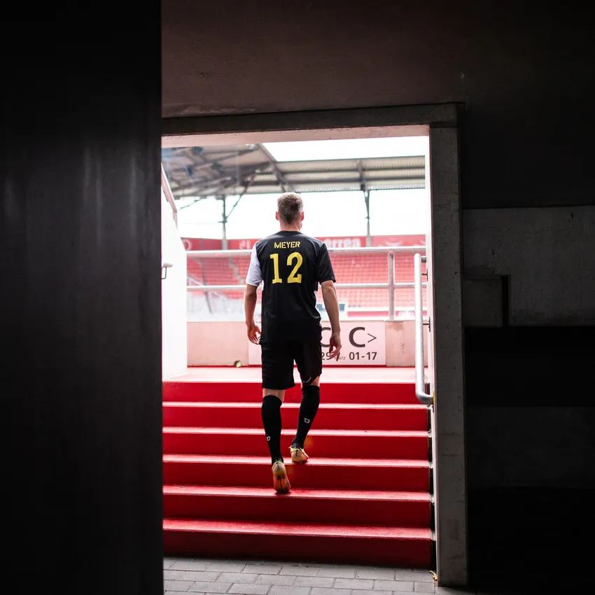 Un calciatore con una maglietta nera con il numero 12 e dettagli gialli, che scende le scale verso uno stadio.