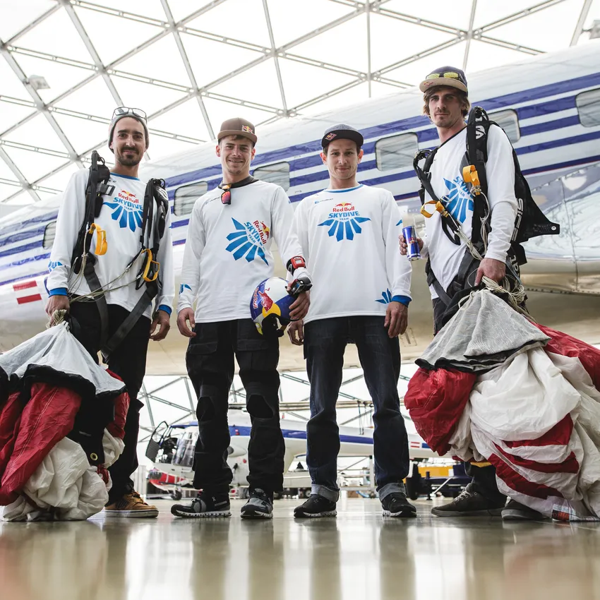 Quattro persone in abbigliamento sportivo si trovano in un hangar moderno, pronte per un salto con il paracadute.