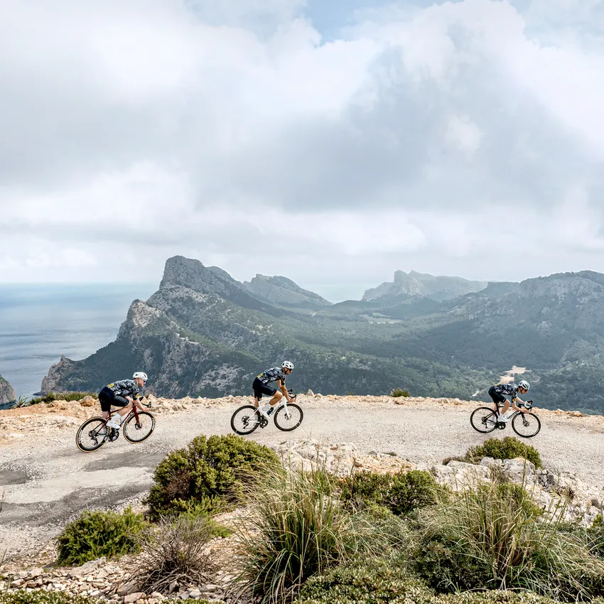 Tre ciclisti in abbigliamento sportivo pedalano su una strada tortuosa con un paesaggio montano e il mare sullo sfondo.