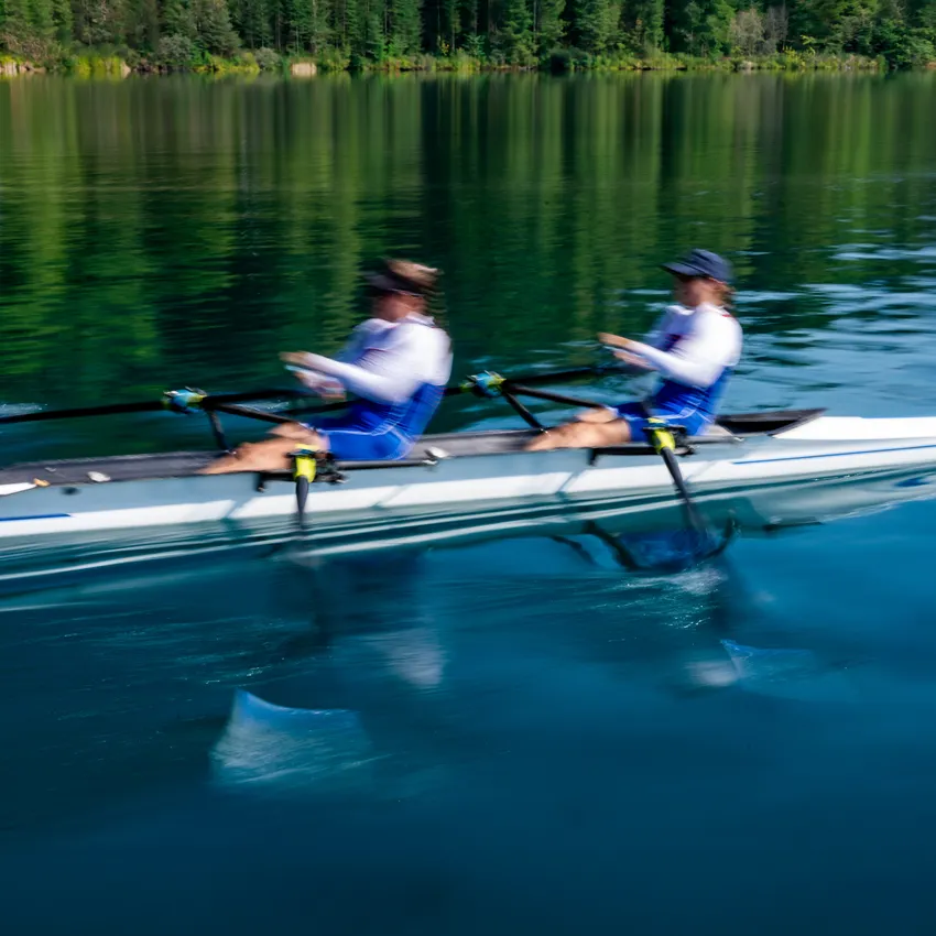 Due rematori in abbigliamento sportivo blu che eseguono colpi potenti su un lago tranquillo.
