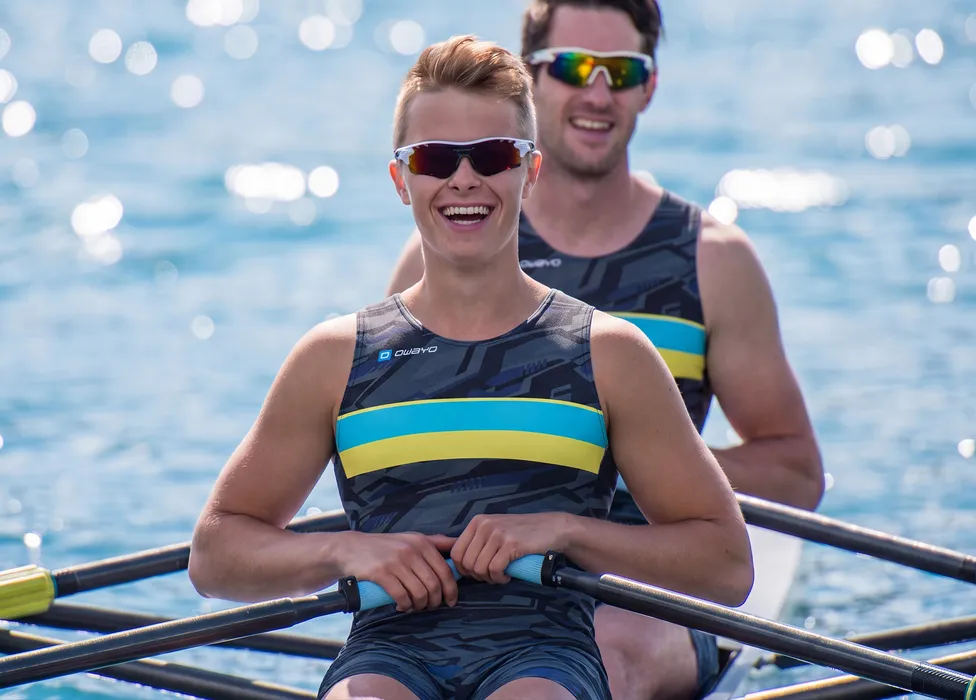 Due rematori in tute sportive a motivi con strisce blu e gialle, sorridendo su un corpo d'acqua.