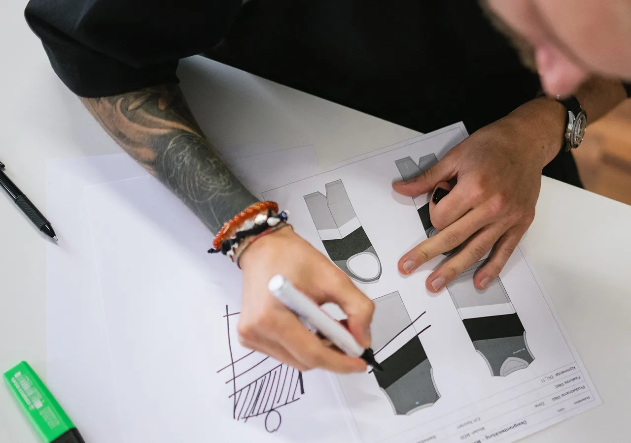 Una persona con un tatuaggio sul braccio disegna con un pennarello bianco su un foglio di carta con diversi progetti di abbigliamento sportivo.