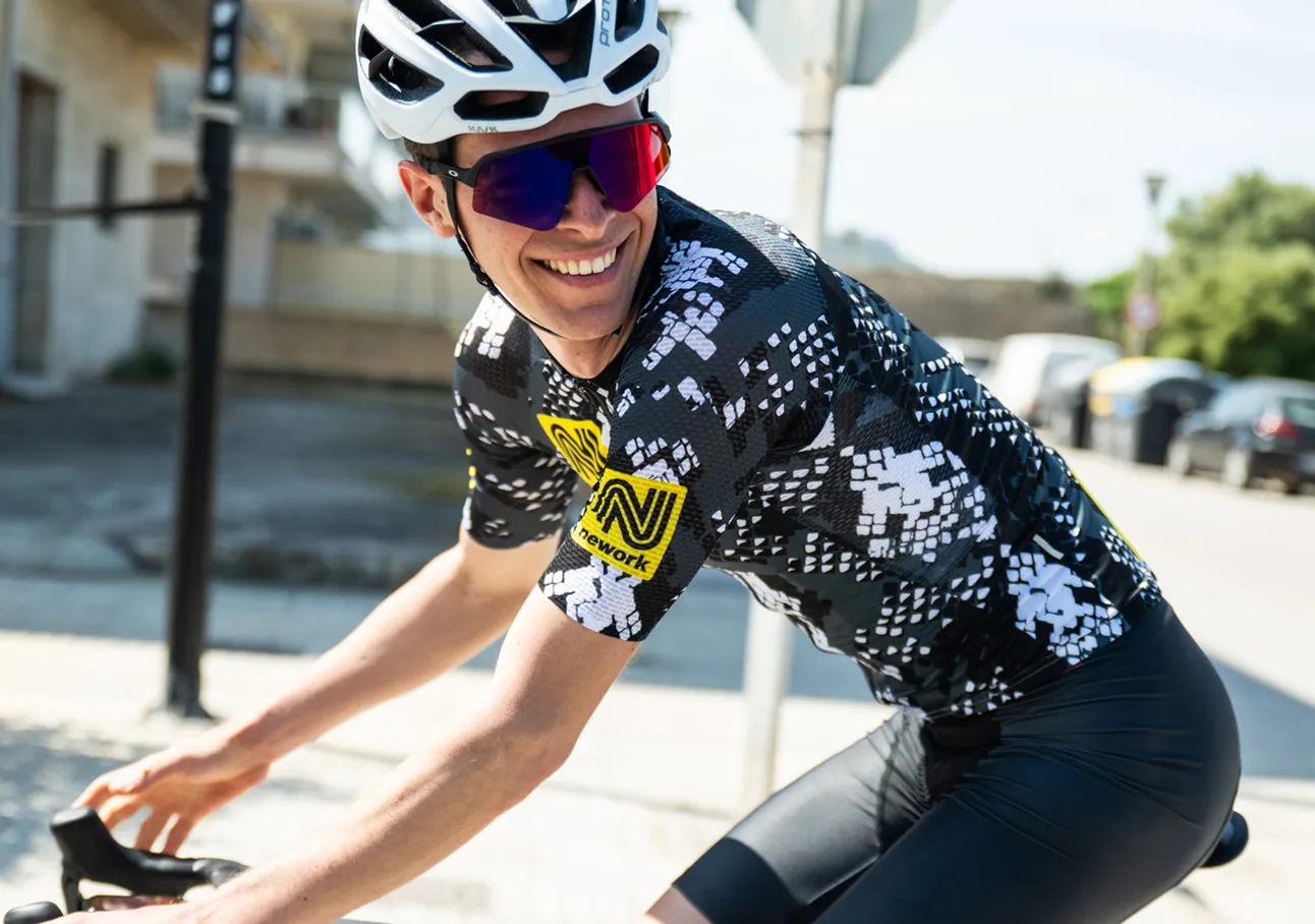 Un ciclista indossa una maglia da ciclismo nera e bianca con un logo sponsor giallo, che pedala per una strada.