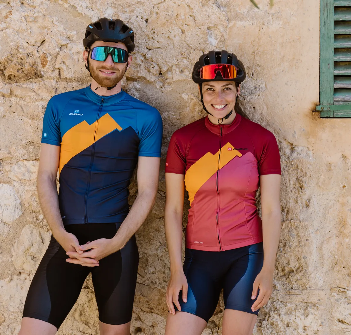 Un uomo e una donna con maglie da ciclismo personalizzate di owayo, in piedi davanti a un muro di pietra.