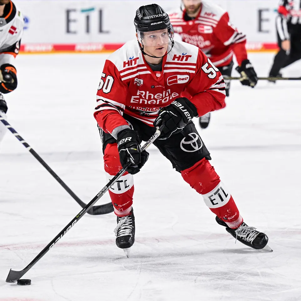 Un giocatore di hockey su ghiaccio con una maglia rossa con il numero 50 e la scritta 'Rhein Energie', in azione sul ghiaccio, controllando un disco con la sua stecca.