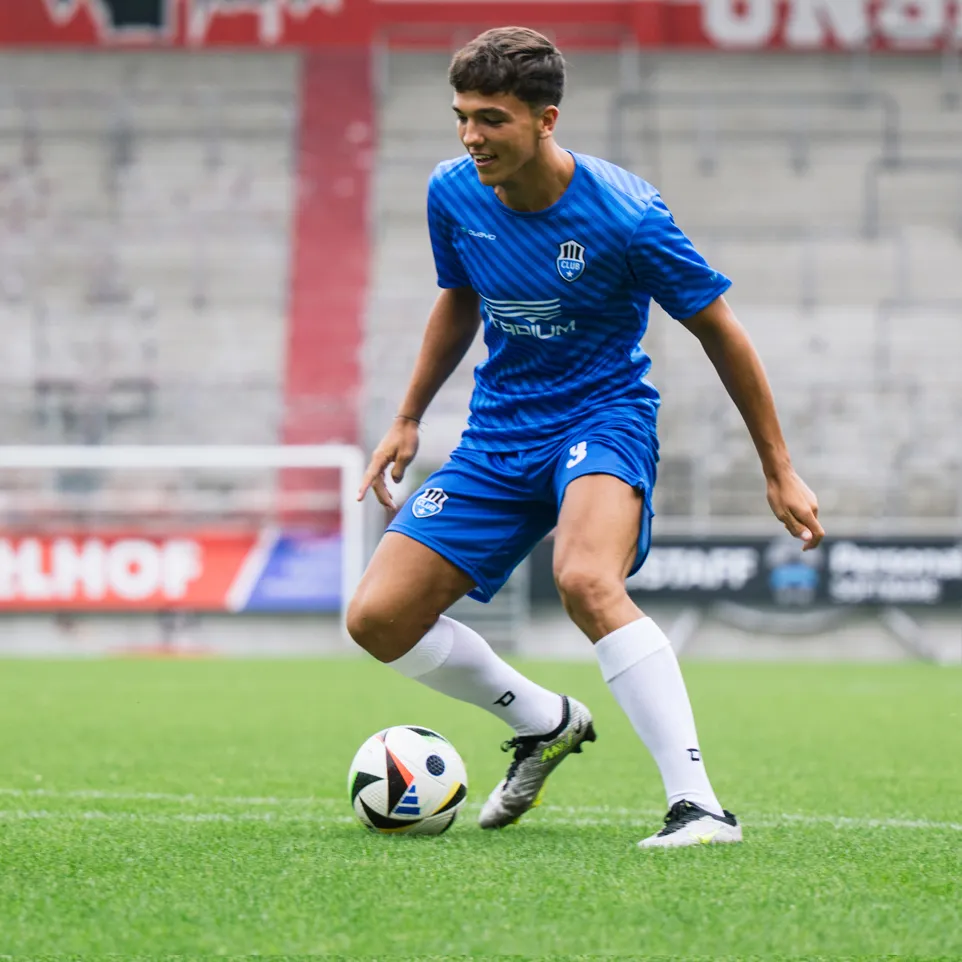 Un giovane calciatore in una maglia blu e pantaloncini blu con un logo sul petto e il numero 3 sui pantaloncini dribbla un pallone da calcio su un campo di erba verde.