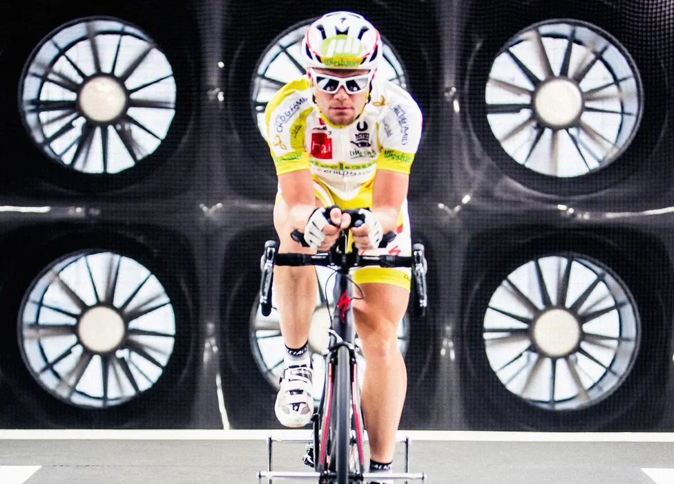 Un ciclista in una maglia gialla e bianca e casco pedala su una bici da allenamento davanti a uno sfondo con grandi ventilatori.
