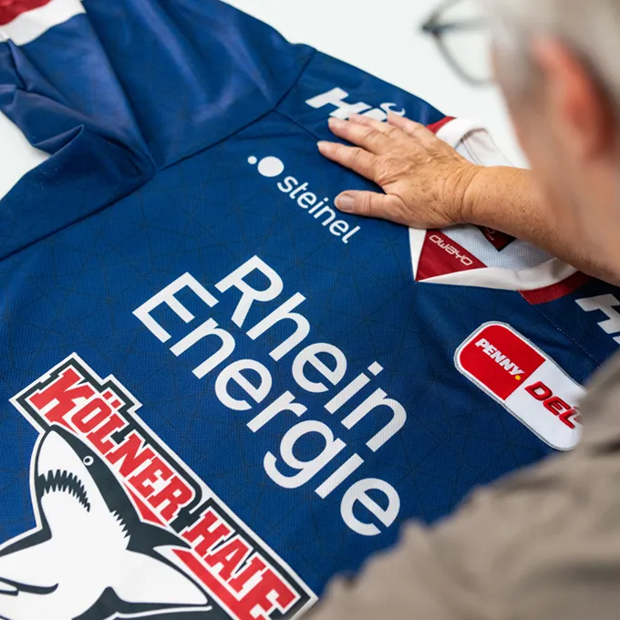 Una persona sta esaminando una maglia sportiva blu personalizzata di owayo con vari loghi di sponsor, tra cui 'Rhein Energie' e 'Kölner Haie'.