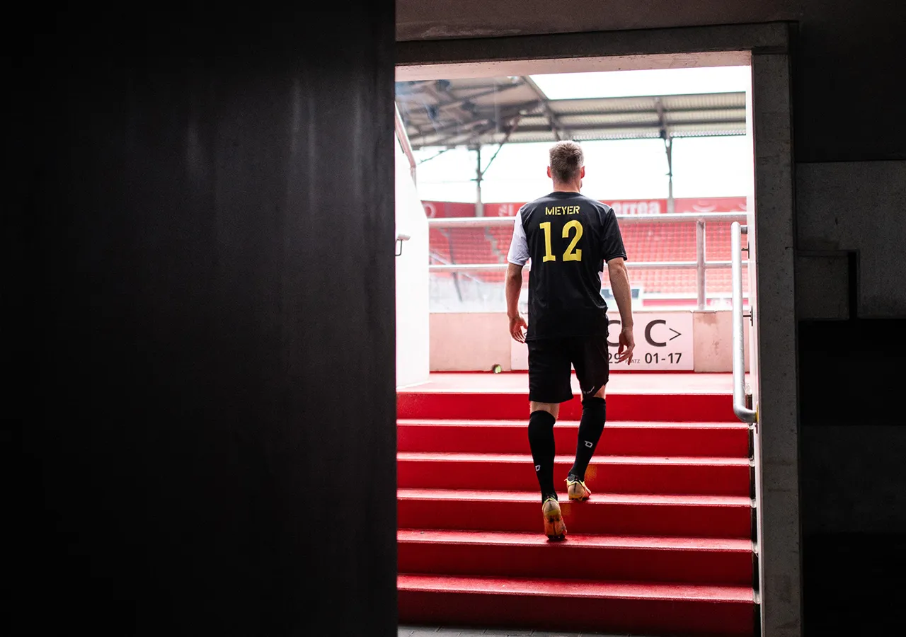 Un calciatore con il nome Meyer e il numero 12 sulla maglia sale una scala rossa verso uno stadio.