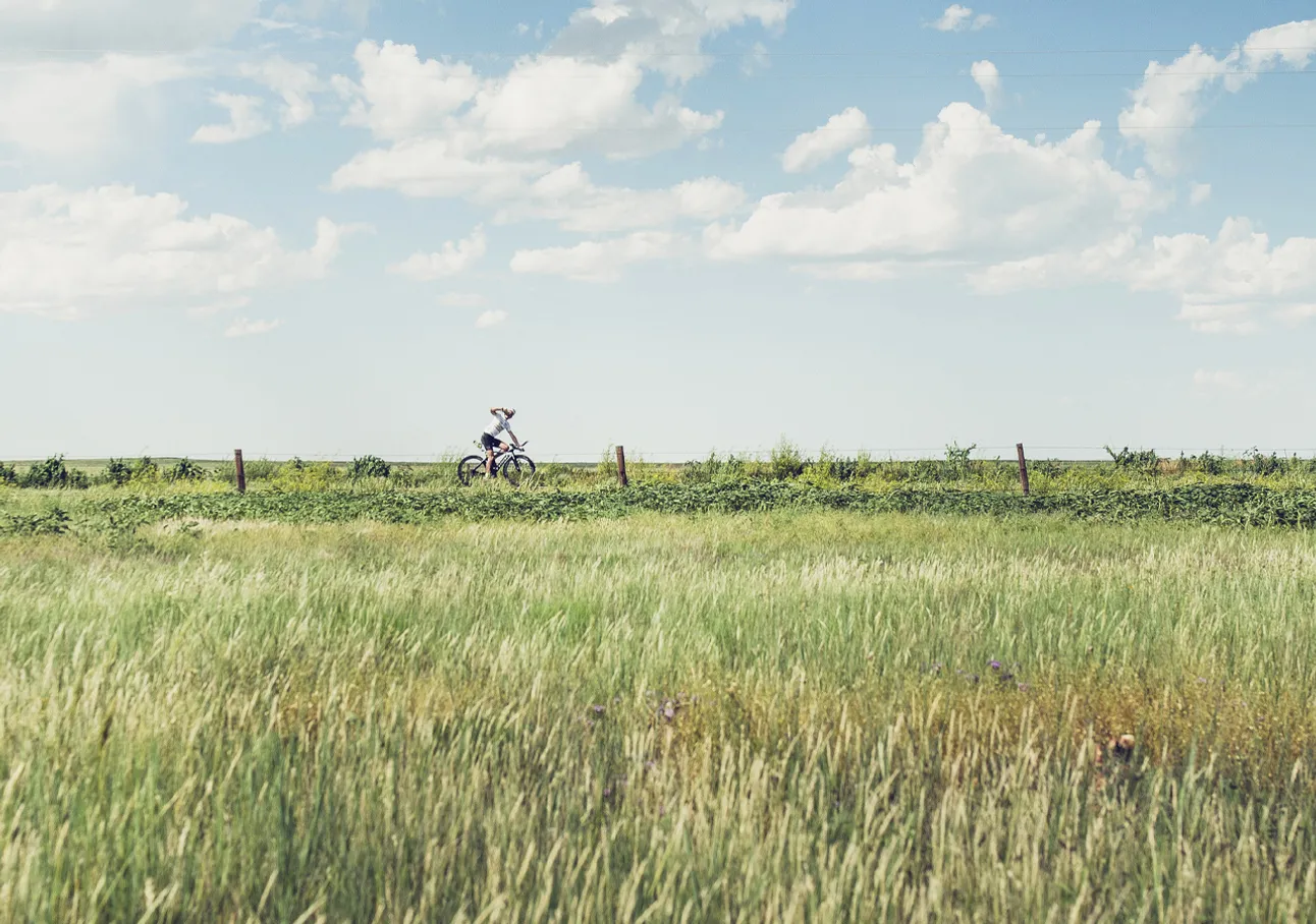 Una persona pedala in bicicletta attraverso un campo verde sotto un cielo blu con nuvole bianche.