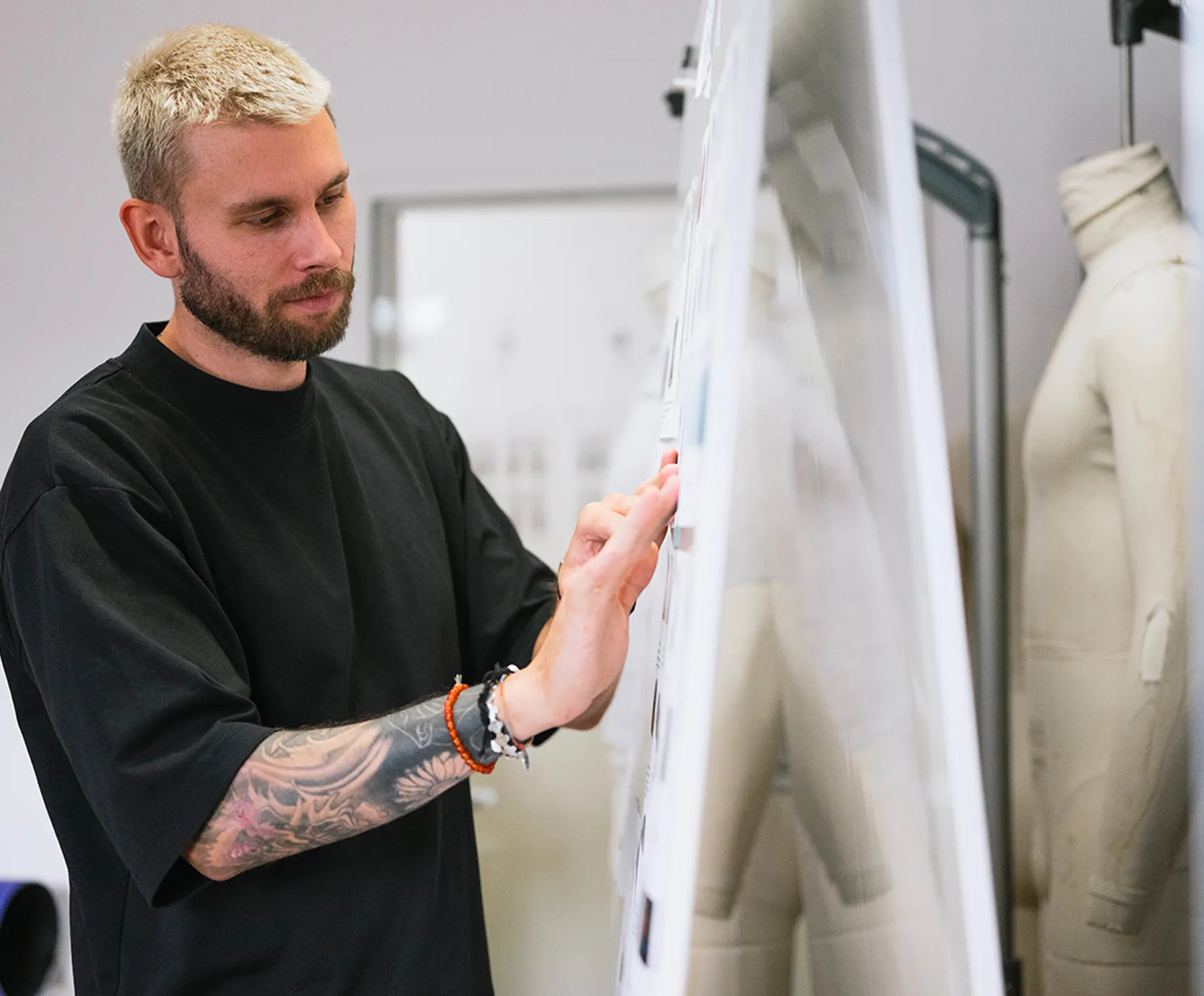 Un uomo con capelli biondi e un tatuaggio sul braccio osserva attentamente dei disegni su una bacheca in uno studio di design, mentre una manichino è visibile sullo sfondo.
